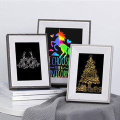 Three framed artworks are displayed on a table. The left frame shows a white stag on a black background. The middle frame depicts a colorful unicorn with the text "I CHOOSE TO BE A UNICORN." The right frame, crafted with CrealityFalcon's A4 Gold Silver Scratch Paper for Falcon Laser Engraving - Pack of 40pcs, features an ornate Christmas tree on a black background.