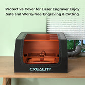 A black CrealityFalcon protective cover for a CR-Laser Falcon 10W Engraver Complete Crafting Engraver Set sits on a wooden table. The cover features an orange-tinted viewing window and a hose attachment at the back. The background shows partially open white blinds with some greenery outside. Text above reads, 