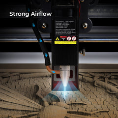 A close-up of a laser engraver in action with the text "Strong Airflow" in the top left corner. The machine is shown engraving a wooden surface with arrows indicating powerful airflow directing dust away from the laser, ensuring clean surfaces. Safety and technical specs are visible on the device. Pictured is the CrealityFalcon Air Assist for CR-Laser Falcon 7.5W and 10W Engraver.